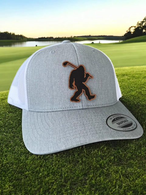 Bigfoot Patch Golf Hat With Sasquatch Holding A Golf Club Heather White Trucker Snapback For Men And Women on grass by lake