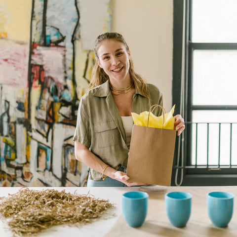 Person holding Small Brown Kraft Paper Bags With Handles 100 Pack 5.25 By 3.25 By 8 Inch Bulk Gift Bags For Crafts Shopping Party Favors Bakery Retail Handmade Treats, filled with yellow tissue paper.