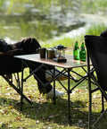 Foldable Aluminum Camping Table With Carry Bag Ultralight Roll Up Design Adjustable Height Indoor Outdoor Use Sturdy Easy Set Up For Camping Picnics Backyard BBQ Patio Wood Grain Finish used outdoors with drinks and lantern placed.