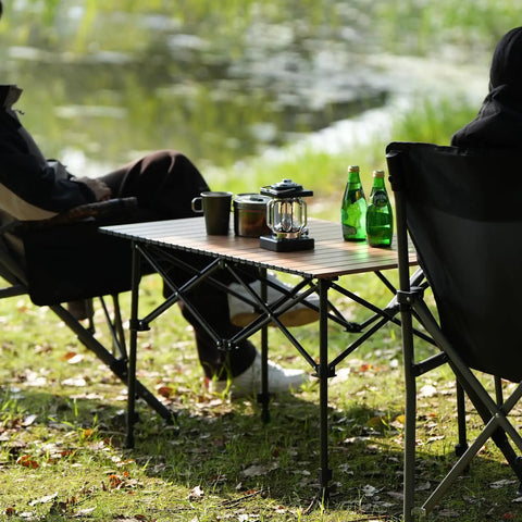 Foldable Aluminum Camping Table With Carry Bag Ultralight Roll Up Design Adjustable Height Indoor Outdoor Use Sturdy Easy Set Up For Camping Picnics Backyard BBQ Patio Wood Grain Finish used outdoors with drinks and lantern placed.