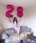 Person holding hot pink number 2 and number 8 balloons on a floral sofa in a living room setting, indoor decor.