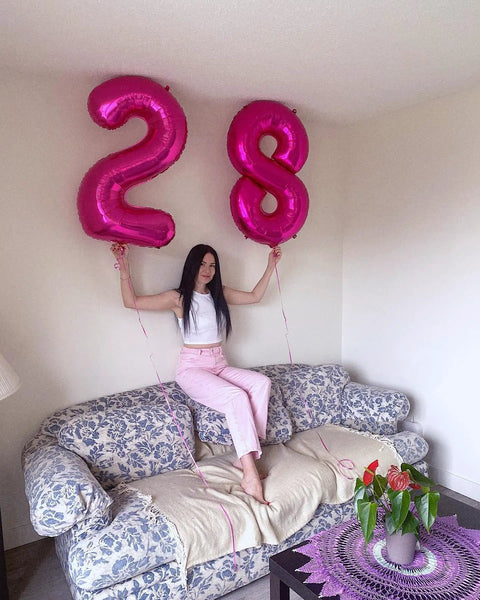 Person holding hot pink number 2 and number 8 balloons on a floral sofa in a living room setting, indoor decor.