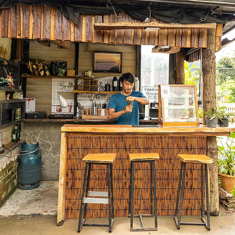 Small outdoor bar with wooden counter and stools, bamboo decor, man preparing drink.