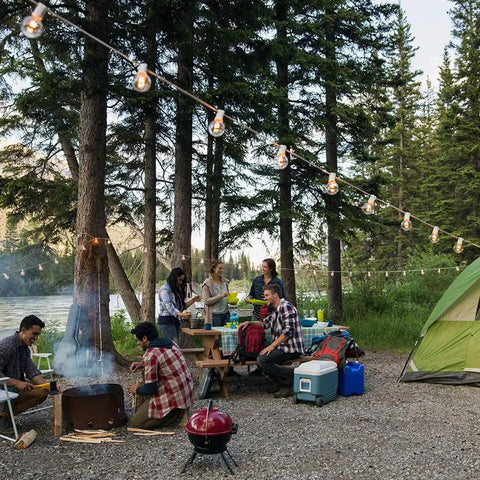 97 Ft White Outdoor String Lights With 49 Shatterproof G40 LED Bulbs hanging above a campfire gathering in a forest setting.
