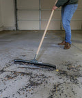 Tough As Nails Twenty Four Inch Curved Steel Floor Squeegee With Handle Heavy Duty Galvanized Scraper clearing debris from garage floor.
