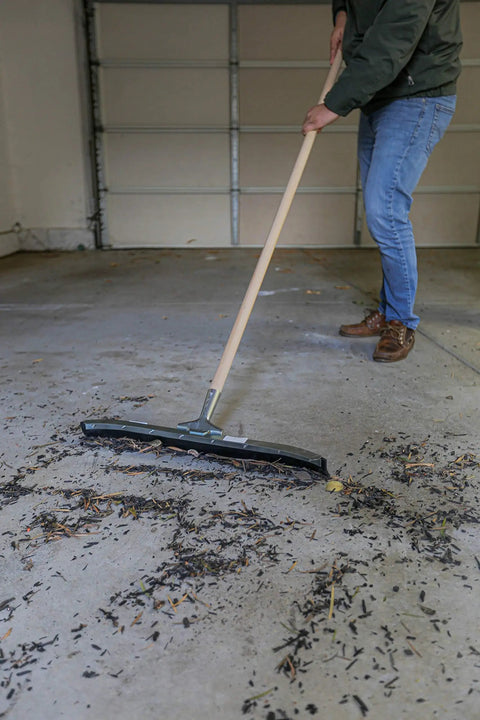 Tough As Nails Twenty Four Inch Curved Steel Floor Squeegee With Handle Heavy Duty Galvanized Scraper clearing debris from garage floor.