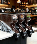 Commercial 4 Pack Coffee Dispenser Straight Up Heavy Duty Countertop Storage Jars For Beans Grains Snacks And Candy Airtight Clear Bulk Food Canisters, displaying coffee beans in a cafe.
