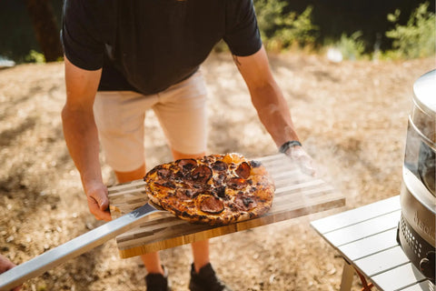 Person using pizza peel to serve pizza next to outdoor oven