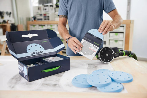 Person holding Granat D125 GR sanding disc near box with VOC varnish discs and sander on table