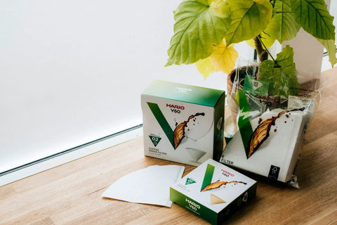 V60 Size 02 Cone Coffee Filters Natural 100 Count Box For Pour Over Brews next to green plant on wooden counter