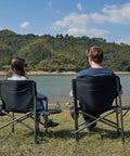 Two people sitting on Ultra Lite Aluminum Oversized Camping Chair With Side Table And Detachable Pocket Portable Directors Chair For Outdoor Camping Lawn And Picnic Supports 400 Lbs Black by a lake
