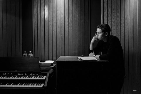 Person sitting beside an organ in a wooden-paneled room, deep in thought, with a notepad on the table.