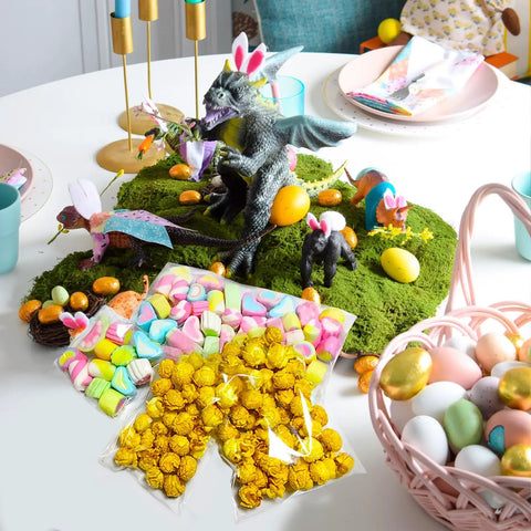Easter-themed table with dragon figurines, moss, and baskets of colored eggs, using Clear 4x4 Inch Resealable Cookie Bags for packaging treats.