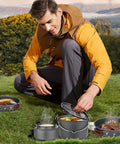 Man using 22 Piece Camping Cookware Mess Kit For Four With Hanging Pot Pan Kettle Base Dishes Cups Spoons Forks And Carry Bags Outdoor Cooking Set For Camping Hiking And Picnics in a scenic outdoor setting.