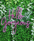 Hello Gorgeous Pink LED Neon Sign For Wall Decor, 21 Inch Neon Light hanging on greenery with vines, acrylic back visible.
