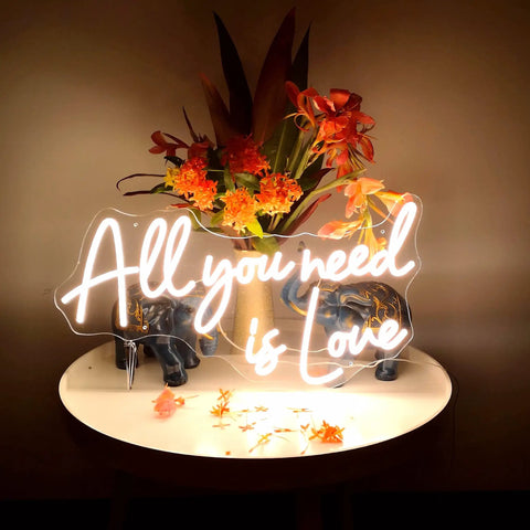 Love Neon Sign LED Neon Light For Wall Decor Warm White Glow on table with flowers, "All you need is Love" text illuminated