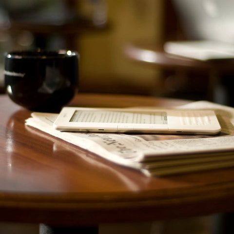 Sleek White Second Gen Ereader Features A Sharp Six Inch Screen And Free 3G Wireless To Pull Down Books Anywhere on a wooden table next to a coffee cup.