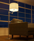Theta Swing Arm Floor Lamp With Brushed Steel Finish, two adjustable arms, drum shade beside a chair in living room corner.