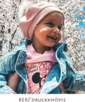 Child wearing pink hat and denim jacket in front of cherry blossom trees, smiling.