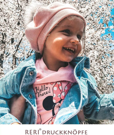 Child wearing pink hat and denim jacket in front of cherry blossom trees, smiling.