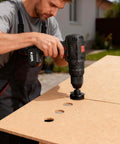 KATA cordless drill with hole saw attachment drilling holes in an OSB board; side view.