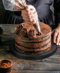 Close-up of a hand piping chocolate frosting on a layered cake using a piping bag, showcasing cake decorating techniques.