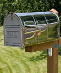Stainless steel mailbox mounted on wooden post in a grassy yard with trees in the background.