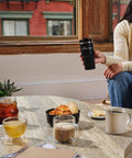 Person holding a black travel mug with assorted beverages and pastries on a table, showcasing an indoor coffee setting.