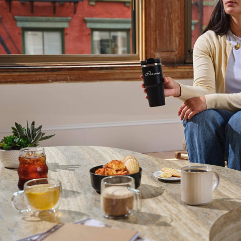 Person holding a black travel mug with assorted beverages and pastries on a table, showcasing an indoor coffee setting.