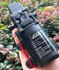 Person holding a 12V DC Fresh Water Pump Diaphragm Self Priming Sprayer Camping Ready For RV Boat Camper Marine Lawn 4.5 L Per Min 110 PSI Adjustable 36W Low Power Consumption With Pressure Switch And 2 Hose Clamps against a garden background.
