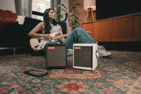 Woman playing guitar near Spark MINI 10W portable guitar amp and Bluetooth speaker with 8-hour battery life on a carpet.