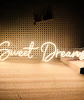 Lit Neon Sign Wall Art Decor 25x6.4 Inches Real LED Neon Light For Bedroom Office Dorm Farmhouse Vibe Aesthetic Room Decor Valentine Party Hanging Sign displaying 'Sweet Dreams' glowing on a table with books