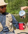 Person pouring coffee from a stainless steel percolator into a mug outdoors, showcasing Stainless Steel Percolator Coffee Pot With Silicone Handle For Camping And Travel Stove Safe 6 Cup Campfire Brew Glacier Stainless Steel Design For Rich Full Bodied Flavor On The Go.