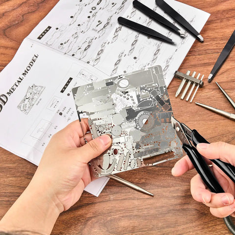 Hands using 12 Piece Metal Model Tool Set For DIY 3D Puzzles Includes Long Edge Bending Tools Tab Twisting Tool Cone Bend Aids Clipper And Four Metal Tweezers For Precise Assembly with metal sheet and instructions on wooden table.