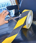 Person cutting yellow and black safety grip tape with scissors diagonal view, focus keyword is No Slip Grip Tape For Indoor Outdoor Use White 1 Inch By 10 Yards All Weather Traction Tape For Stairs Floors Decks Ladders And Equipment With 60 Grit Aluminum Oxide For Maximum Traction. Roll of tape.
