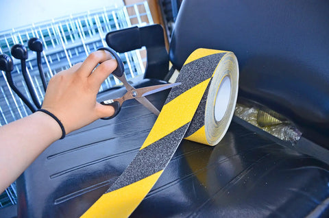 Person cutting yellow and black safety grip tape with scissors diagonal view, focus keyword is No Slip Grip Tape For Indoor Outdoor Use White 1 Inch By 10 Yards All Weather Traction Tape For Stairs Floors Decks Ladders And Equipment With 60 Grit Aluminum Oxide For Maximum Traction. Roll of tape.