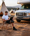 Man using a laptop in a folding chair next to a camper van, surrounded by tents while camping.