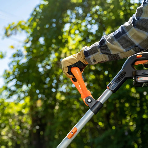 Close-up of a hand gripping the handle of a 20V Cordless Garden Tiller Cultivator with 7.5 Inch Width, set against greenery.