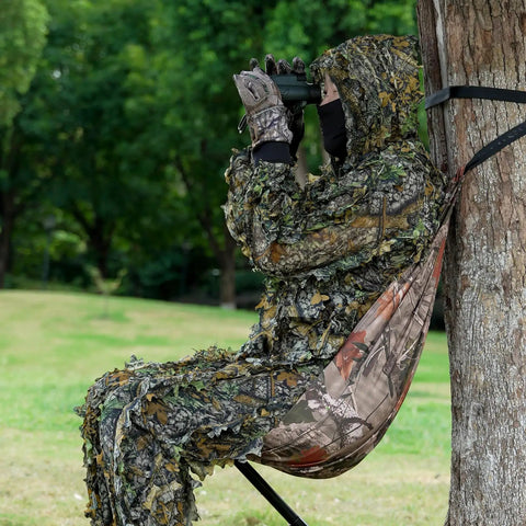 Person seated in Camo Tree Hammock Chair Portable Hunting Stool For Outdoors attached to a tree in a camouflage outfit, using binoculars.