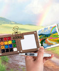 Mini Watercolor Travel Kit With Wooden Palette And Copper Clip Includes 24 Watercolors 6 Brushes And 2 Sketchbooks For Beginners And Painting Enthusiasts On The Go displayed open with painting scenery background