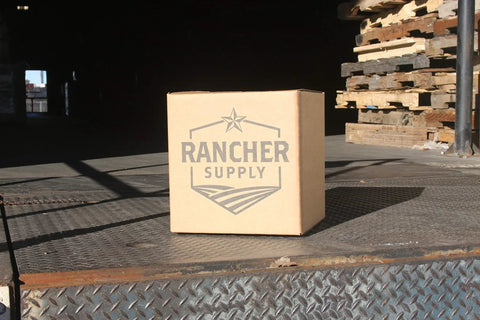 Cardboard box labeled 'Rancher Supply' on a metal platform near stacked pallets in a warehouse