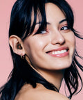 Woman wearing Chill Quiet Ear Plugs For Noise Reduction With Ultra Soft Silicone Comfort Reusable Sleep Protection Eight Tip Sizes XS S M L 24dB SNR 14 NRR Noise Cancelling In Black Plus Four Color Options, smiling on pink background.