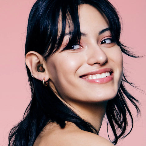 Woman wearing Chill Quiet Ear Plugs For Noise Reduction With Ultra Soft Silicone Comfort Reusable Sleep Protection Eight Tip Sizes XS S M L 24dB SNR 14 NRR Noise Cancelling In Black Plus Four Color Options, smiling on pink background.