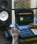 Person using laptop with music production software, next to HS7 7-Inch Powered Studio Monitor Pair With Bi-Amp System, 43 Hz To 30 kHz Range.