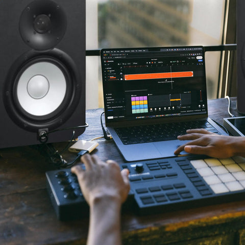 Person using laptop with music production software, next to HS7 7-Inch Powered Studio Monitor Pair With Bi-Amp System, 43 Hz To 30 kHz Range.