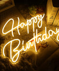 "Happy Birthday" neon sign glowing warmly on tabletop next to laptop and books, ideal for party decor.