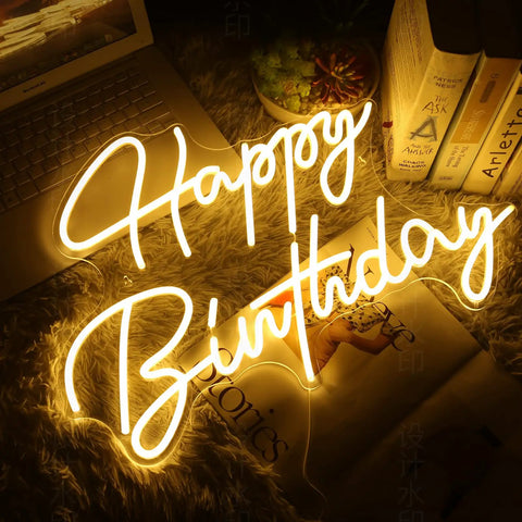 "Happy Birthday" neon sign glowing warmly on tabletop next to laptop and books, ideal for party decor.