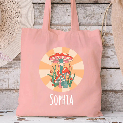 Canvas tote bag with mushroom design and 'Sophia' text, displayed against a rustic wooden wall with straw hats.