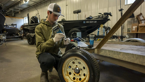 Quicksilver 2-4-C marine grease 8 oz tube being applied to a trailer wheel; blue packaging visible.