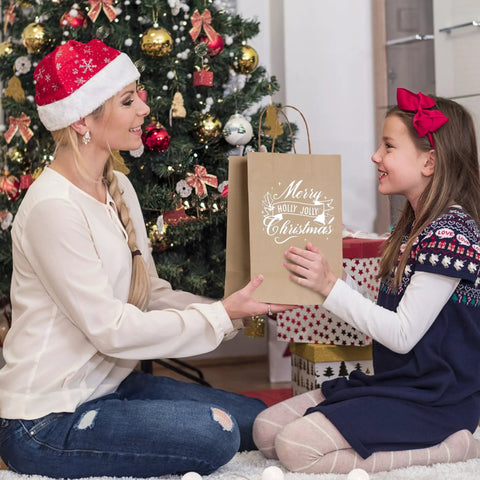 Mother and daughter exchanging 100 Pack Kraft Paper Gift Bags With Handles, Medium Brown, Christmas scene, tree in background.